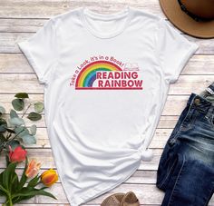 Step into nostalgia with the Reading Rainbow Shirt, a timeless piece that merges classic charm with modern comfort. Crafted from premium cotton, this shirt embodies casual elegance and irresistible softness. The design features a subtle nod to the beloved Reading Rainbow show, making it a must-have for fans and fashion enthusiasts alike. Whether worn solo or layered for a trendy look, this tee effortlessly elevates any outfit. Embrace the joy of reading and style with the Reading Rainbow Shirt, a versatile and iconic addition to your wardrobe. Let this shirt be a statement piece that celebrates literature and showcases your unique sense of fashion effortlessly. The solid color Unisex Tees are super soft ring-spun cotton...it's sure to be your new favorite tee (heather tees are a soft cotto Book Shirt, Librarian Shirt, Reading Rainbow, Rainbow Fashion, Rainbow Shirt, Book Shirts, Text Design, Book Lovers Gifts, Casual Elegance