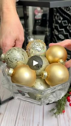 two hands holding a glass bowl filled with gold and silver christmas ornaments on top of a wooden table