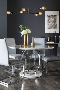 a dining room table with white chairs and gold vases on the top, in front of a gray wall