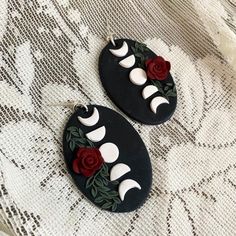 two black oval earrings with red roses and crescent moon designs on the sides, sitting on a lace surface