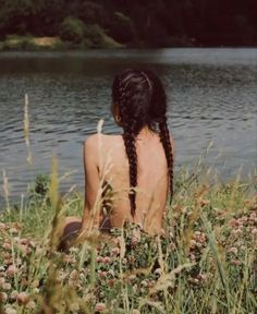 a woman sitting in the grass by a lake with her back turned to the camera