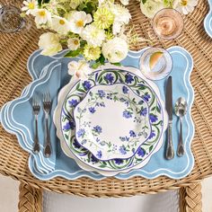 a wicker table with blue and white plates, silverware and flowers on it