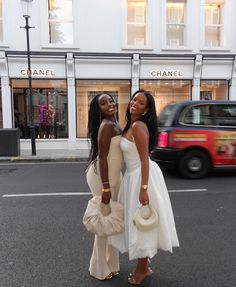 two beautiful women standing next to each other in front of a tall building on a city street