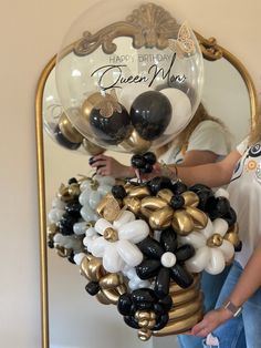 a woman standing in front of a mirror holding a bunch of black, white and gold balloons