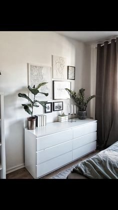 a white dresser sitting next to a window with pictures on the wall and plants in vases