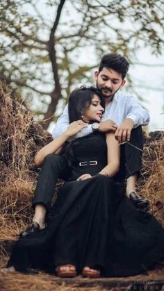 a man and woman are sitting on the ground in front of some trees with their arms around each other