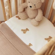 a teddy bear sitting on top of a crib next to a white blanket and pillow