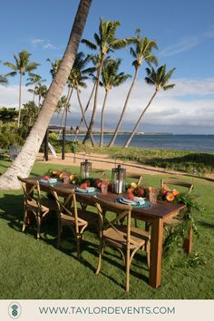 an outdoor dining table set up on the lawn with palm trees and ocean in the background