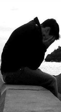 a man sitting on top of a cement wall next to the ocean with his head in his hands