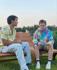 two young men sitting on wooden benches in the grass looking at each other and smiling