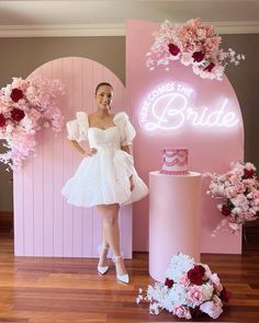 a woman standing in front of a pink backdrop with flowers and a cake on it