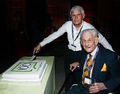 an older man cutting into a cake with a knife and fork while another person looks on