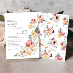 two wedding cards with flowers on them sitting next to some white and pink roses in the background