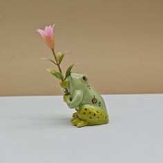 a frog figurine holding a pink flower in it's mouth