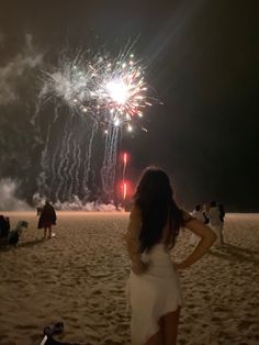 a woman standing on top of a sandy beach next to a firework display in the sky