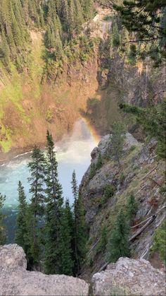 a rainbow shines in the sky over a river and trees on top of a cliff