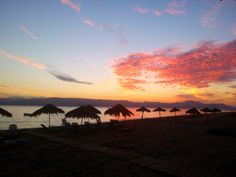 the sun is setting on the beach with umbrellas