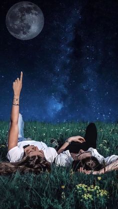 two people laying in the grass under a full moon and stars filled sky, with their hands up