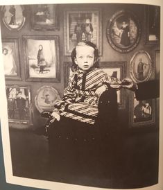 an old black and white photo of a young boy sitting in a chair with pictures on the wall behind him