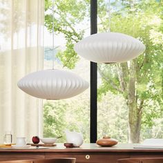 two white lamps hanging over a dining table next to a window with trees in the background