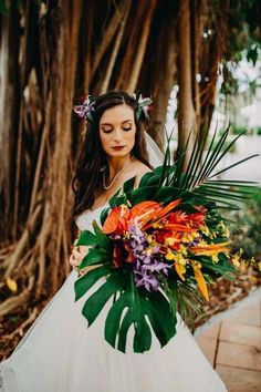 a woman in a wedding dress holding a bouquet of flowers and greenery with trees behind her