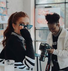 two women sitting at a table with drinks and camera equipment in front of their faces