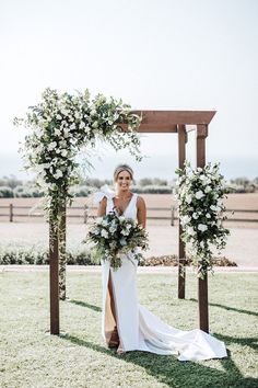 a woman in a white dress is standing under an arch with flowers and greenery