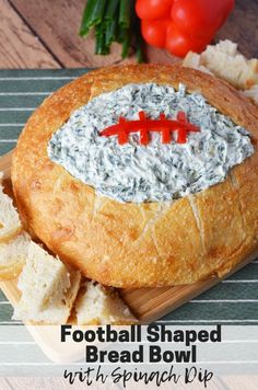 a football shaped bread bowl with spinach dip in it on a cutting board next to crackers and tomatoes