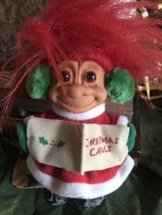 a troll doll with red hair and green eyes holding a christmas carol's sign