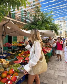 a woman standing in front of a market filled with lots of fresh fruits and vegetables