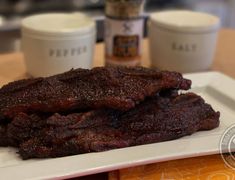 two pieces of steak on a white plate next to some coffee cups and sauces