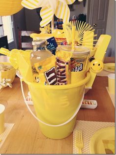 a yellow bucket filled with lots of food on top of a wooden table next to utensils