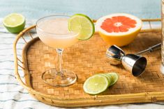 a drink in a glass on a tray with limes and grapefruit next to it