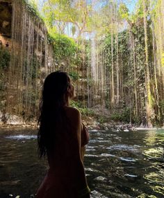 a woman standing in the water looking up at a waterfall with trees hanging from it