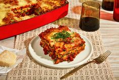 a white plate topped with lasagna next to a red casserole dish