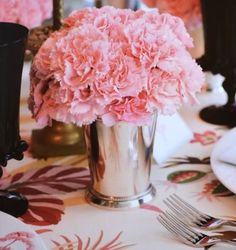 pink carnations are in a silver vase on a table with plates and utensils