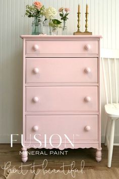 a pink dresser sitting next to a white chair and vase with flowers on top of it