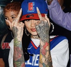 a woman with tattoos covering her eyes and making the peace sign while standing in front of a group of people