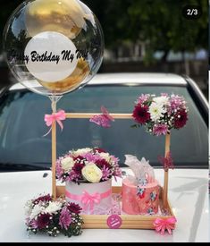 a birthday cake and balloons on the hood of a car with flowers in front of it