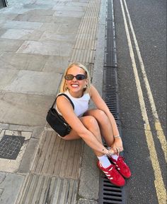 a woman sitting on the side of a road with her legs crossed and wearing red sneakers