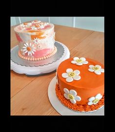 two cakes sitting on top of wooden table next to each other, one with orange frosting and the other decorated with white flowers