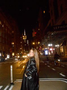 a woman standing on the side of a road at night with buildings in the background