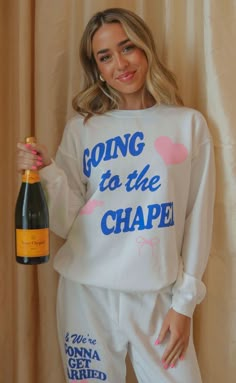 a woman in pajamas holding a bottle of wine and smiling at the camera while wearing a sweatshirt that says going to the chapel