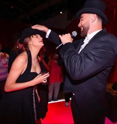 a man in a black suit and hat sings into a microphone while a woman in a black dress stands next to him