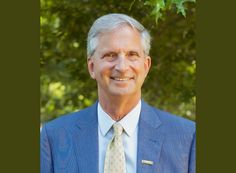 an older man wearing a blue suit and yellow tie smiling at the camera with trees in the background