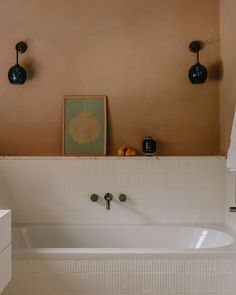 a white bath tub sitting next to a sink under two black light fixture lights and a painting on the wall