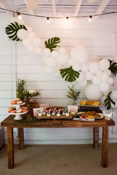 a wooden table topped with lots of food and white balloons hanging from the ceiling above it