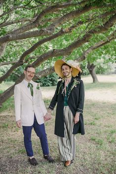 two people standing next to each other in front of some trees holding hands and looking at the camera