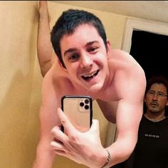 a shirtless man taking a selfie in front of his cell phone while another man looks on