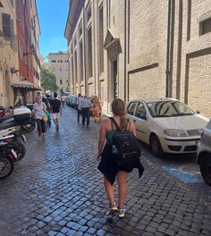 a woman walking down a cobblestone street next to parked cars and motor scooters